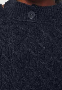 Pull en maille câble bleu marine foncé avec un col rond et un seul bouton foncé au niveau de l'encolure.