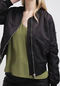 Svart bomberjacka med dragkedja fram, ribbade muddar och krage. Buren över en olivgrön, V-ringad blus, vilket skapar en lager-på-lager-look.