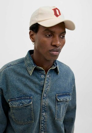 Beige cap with red 'U' logo, worn with a blue denim shirt featuring two chest pockets, textured fabric, and snap buttons.