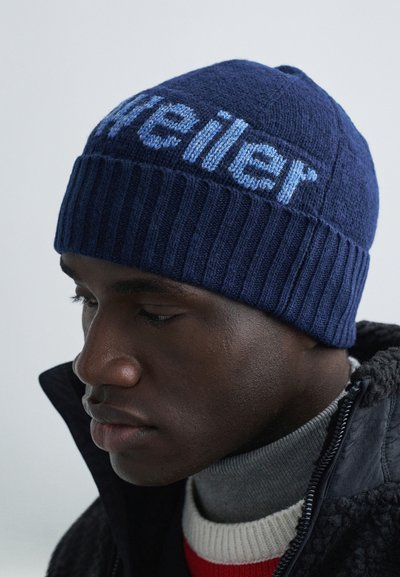Mornarskoplava plava pletena kapa s rebrastom teksturom i svijetloplavim natpisom. Ima zaobljeni vrh i uski kroj, prikladna za zimsko nošenje.