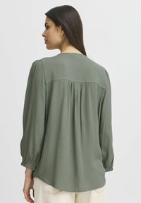 Blusa verde, con texture, dal collo rotondo, maniche a sbuffo e un dettaglio plissettato sul retro. Il materiale sembra leggero e informale.
