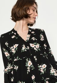 Blusa preta com padrão floral, decote em V, cintura franzida e mangas com babados. Apresenta padrões florais em rosa e verde sobre um tecido leve.