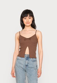 Top crop castanho com nervuras, apresentando alças ajustáveis, um detalhe em laço na frente e um corte na cintura, combinado com jeans azul claro.