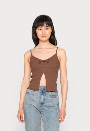 Top crop castanho com nervuras, apresentando alças ajustáveis, um detalhe em laço na frente e um corte na cintura, combinado com jeans azul claro.