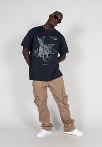 Mørkeblå bomulds t-shirt med grafisk tryk, der viser en figur og ordet "SAINT." Sæt sammen med beige cargo-bukser og hvide sneakers.