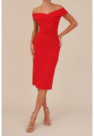 Vrouw met een nauwsluitende, rood kruisjes-middenlange jurk met een afvallende halslijn, gecombineerd met beige enkelsandalen met enkelbandjes en gouden armbanden.