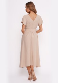 Beiges Midi-Kleid mit kurzen Ärmeln, V-Ausschnitt im Rücken, Gürtel in der Taille und glatter Textur. Getragen mit nuden Riemchen-Pumps.