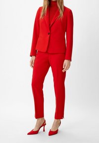 Blazer ajusté rouge avec deux poches, associé à un pantalon slim assorti et des escarpins rouges à bride arrière, sur fond blanc.