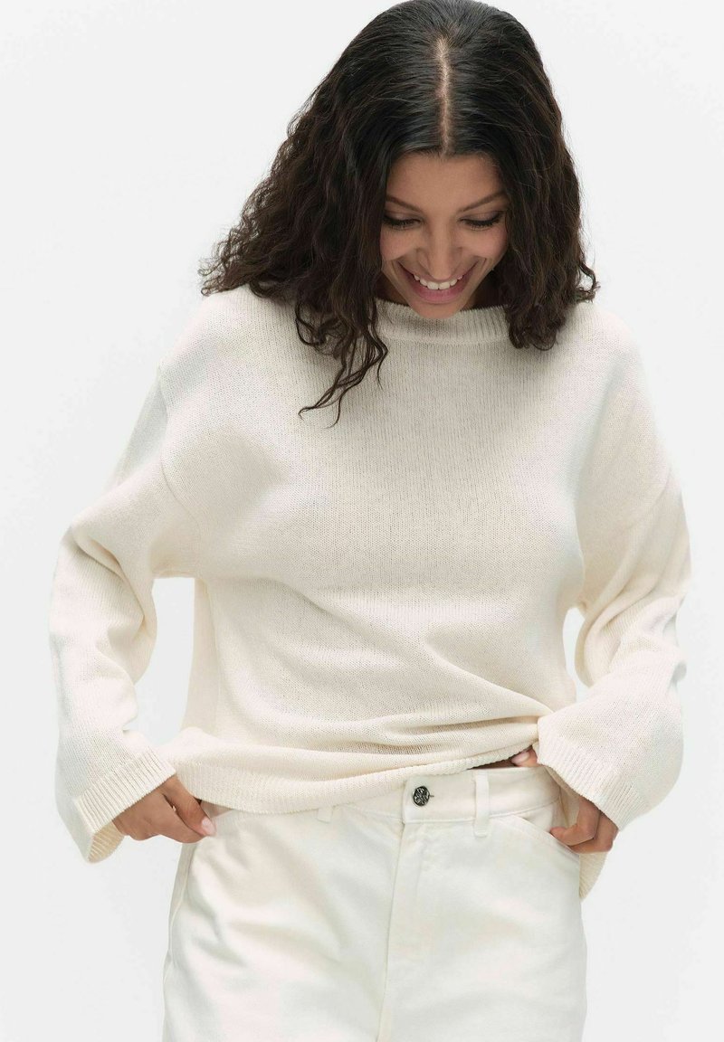 Vrouw met krullend donker haar, een losse crèmekleurige trui en witte broek, glimlachend en naar beneden kijkend terwijl ze haar trui rechttrekt.