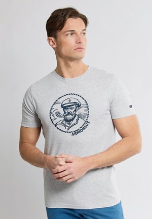 Homme en t-shirt gris avec une illustration de marin bleu marine et le texte « Armor Lux », mains jointes, regardant vers sa gauche sur un fond uni.