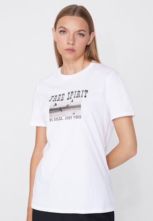Kobieta z prostymi włosami, ubrana w białą koszulkę z czarnym napisem "FREE SPIRIT" oraz "NO RULES, JUST VIBES", stojąca na jednolitym tle.