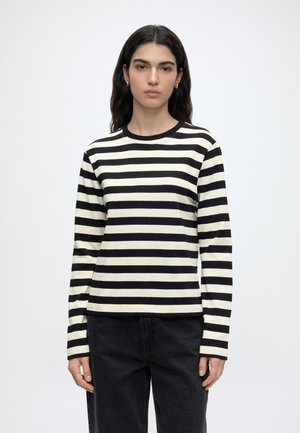Femme aux longs cheveux foncés portant un t-shirt à manches longues à rayures horizontales noires et blanches et un pantalon noir, debout devant un fond uni.