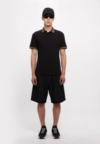 Čierne polo tričko so vzorovaným golierom, vreckom, čierne šortky a čierna čiapka. Slnečné okuliare a čierne tenisky s bielymi textovými akcentmi.