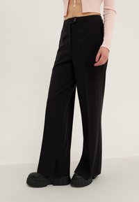 Pantalon noir à jambes larges en tissu lisse, doté d'une fermeture boutonnée à l'avant et de fentes latérales distinctes à l'ourlet.