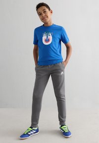 Μπλε βαμβακερό μπλουζάκι με γραφικό της Ιταλίας, γκρι φόρμες και μπλε αθλητικά παπούτσια με φωσφοριζέ πράσινα κορδόνια. Ανεπίσημο σύνολο σχεδιασμένο για άνεση.