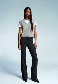 Grauer Strick-Sweater ohne Ärmel über einem weißen Hemd, kombiniert mit schwarzen Streifen-Breitehosen und spitzen schwarzen Schuhen. Einfaches und modernes Outfit.