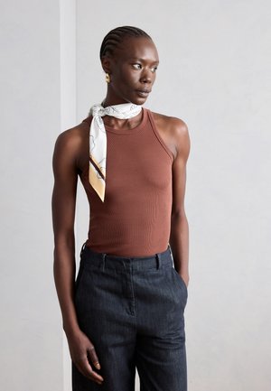 Ženska s pletenicami, ki nosi rjavo zgornjo brez rokavov, temne hlače, vzorčasto belo ruto, zavezano okoli vratu, in zlati uhani.