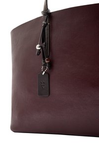 Bordowa skórzana torba typu tote o gładkiej fakturze, zaokrąglonym górnym brzegu, dekoracyjnym chwostem z koralikami oraz czarną skórzaną metką z wytłoczonym wzorem.