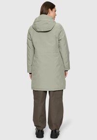 Femme portant un long manteau à capuche gris clair, un pantalon marron foncé et des chaussures noires, se tenant de dos sur un fond clair.