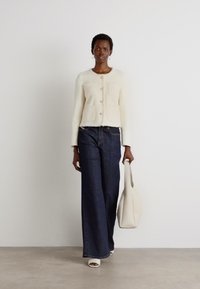 Cream cardigan with square pockets, front buttons, paired with wide-leg dark denim pants. Model holds a light-colored tote bag and wears white shoes.