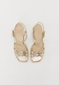 Gullmetalliske slingback sandaler med kvadratisk tå, med kryssede stropper og en glatt tekstur. Sålen er merket som "SUPER MYK."