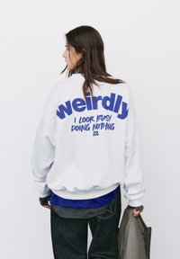Felpa grigia con testo blu "Stranamente sembro occupato a non fare nulla" sulla parte posteriore. Vestibilità ampia, polsini a costine, indossata con una camicia blu sotto.