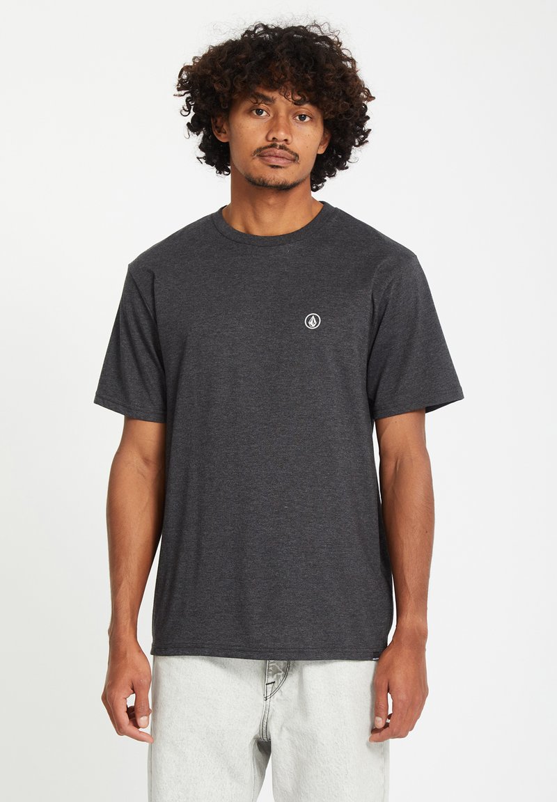 Homme aux cheveux bouclés portant un t-shirt gris foncé à manches courtes et un pantalon de couleur claire, debout devant un fond blanc uni.