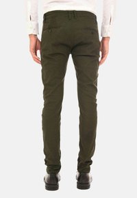Pantaloni attillati di colore verde scuro con un motivo testurizzato, dotati di due tasche posteriori e passanti per cintura, abbinati a scarpe nere.