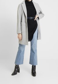 Hellgrauer Mantel mit Reversdesign, kombiniert mit einem schwarzen Rollkragenpullover und blauen Schlagjeans, ergänzt durch schwarze Stiefeletten.