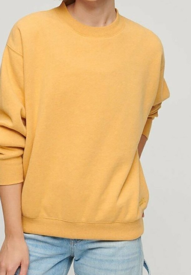 Personne portant un sweat-shirt uni jaune moutarde à manches longues et un jean bleu clair, debout devant un fond neutre.