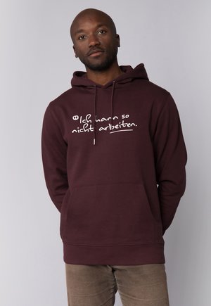 Man met een kastanjebruine hoodie met de Duitse tekst "Ich kann so nicht arbeiten" en beige broek, staand tegen een eenvoudige lichte achtergrond.