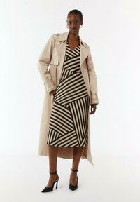 Beige Trenchcoat über einem gestreiften Kleid mit schwarzen und beigen geometrischen Mustern. Stiletto-Absätze und ein Armband betonen das Outfit.
