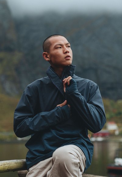 Marinblå vattentät jacka med hög krage, dragkedjestängning och texturerat mönster. Modellen bär ljusbeiga byxor och sitter mot en naturlig bakgrund.