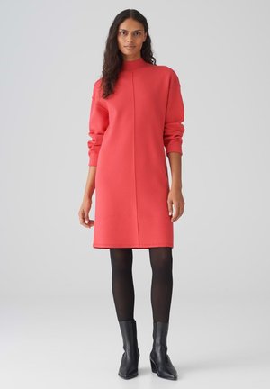 Robe sweat corail avec un col haut, des manches longues et une coupe droite ; assortie à des bottines noires et des collants. Texture de tissu lisse.