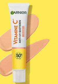 Tube de crème solaire Garnier à la vitamine C, blanche avec un logo vert, présente des accents roses et un label SPF 50+. Échantillons de crème beige chaud à ses côtés sur un fond jaune.