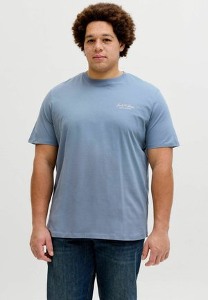 Junger Mann mit lockigem Haar, der ein blaues Jack & Jones T-Shirt und dunkle Jeans trägt, vor einem schlichten weißen Hintergrund stehend.