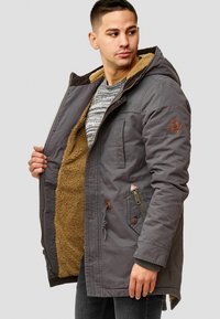 Grijze fleece-gevoerde jas met capuchon, voorzien van een ritssluiting, meerdere zakken en bruine leren accenten op de mouwen en rits.