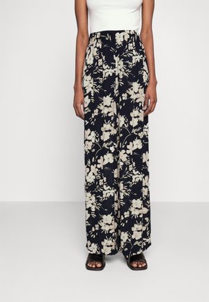 Femme portant un pantalon noir taille haute à jambes larges avec un imprimé floral beige, associé à des sandales noires à bout ouvert et un haut blanc sans manches.