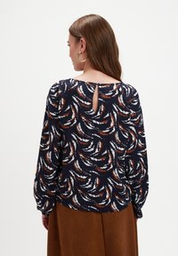 Kobieta z długimi brązowymi włosami, nosząca złote kolczyki w kształcie kół, granatową bluzkę w wzory z dekoltem w kształcie łezki oraz brązową spódnicę.