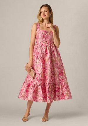 Kvinde i en ærmeløs, lyserød blomster-midi-kjole, der holder en beige clutch, iført sandaler med åbne tæer, stående mod en ensfarvet baggrund.