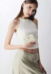 Portefeuille de couleur crème avec un motif floral et des accents dorés, tenu contre un haut gris clair. L'arrière-plan est neutre et minimal.