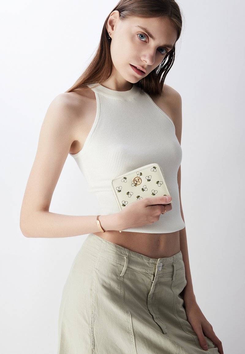 Portefeuille de couleur crème avec un motif floral et des accents dorés, tenu contre un haut gris clair. L'arrière-plan est neutre et minimal.