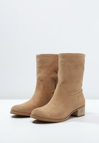 Bottes basses en daim beige avec un embout arrondi, une tige courte, une fermeture éclair latérale et un talon bloc en bois. Texture lisse avec des accents minimaux.