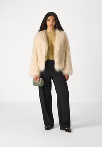Chaqueta de piel sintética crema, blusa amarilla transparente, pantalones de pierna ancha negros y bolso verde de cuentas. La modelo lleva pendientes de aro y zapatos negros.