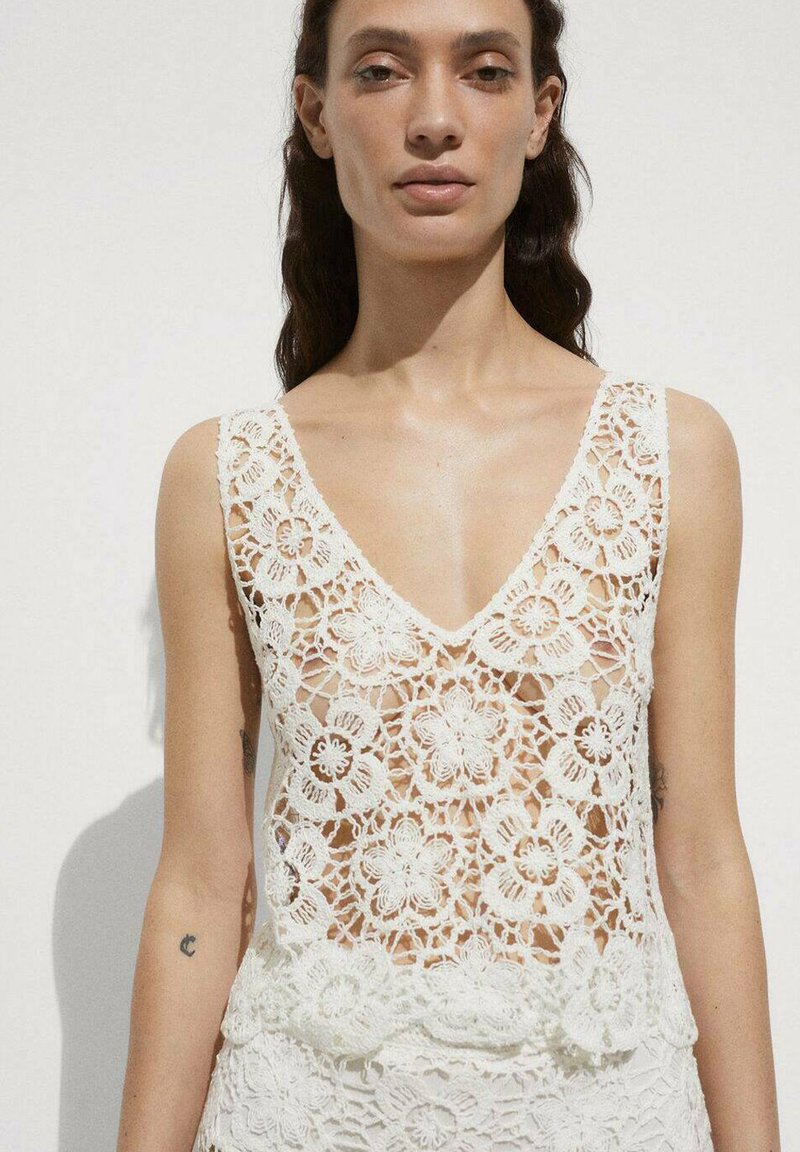 Top din croset alb, cu un model floral, decolteu în V și design transparent. Materialul are un aspect delicat și texturat.
