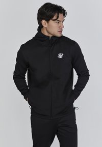 Zwarte zip-up hoodie van zacht materiaal met een aansluitende pasvorm, hoge kraag en een wit logo op de borst. Gladde textuur en minimaal ontwerp.