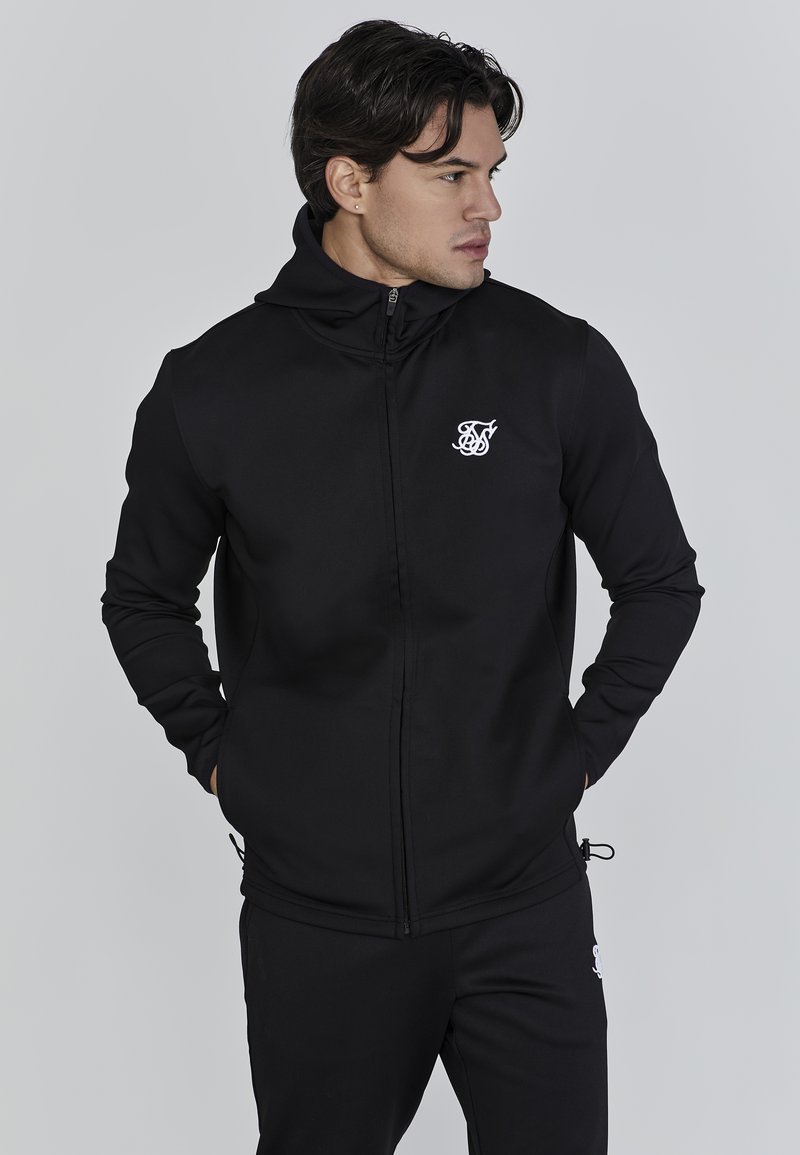 Zwarte zip-up hoodie van zacht materiaal met een aansluitende pasvorm, hoge kraag en een wit logo op de borst. Gladde textuur en minimaal ontwerp.