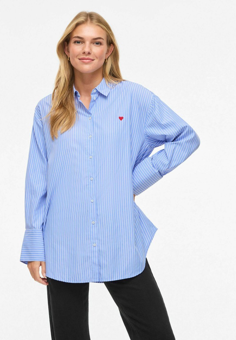 Chemise à rayures bleues avec un bouton-pression et un accent en forme de cœur rouge sur la poitrine, dotée de manches longues et d'une coupe décontractée.