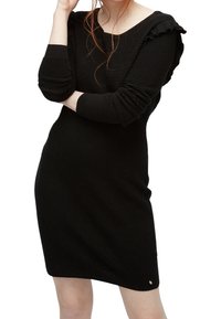 Svart ribbad klänning med långa ärmar, volangdetaljer på axlarna och en figurnära silhuett. Den strukturerade tyget ger en subtil djup.