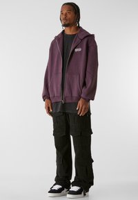 Sudadera morada con cremallera y grandes bolsillos, llevada sobre una camiseta negra; combinada con pantalones cargo negros con múltiples bolsillos y un ajuste relajado.